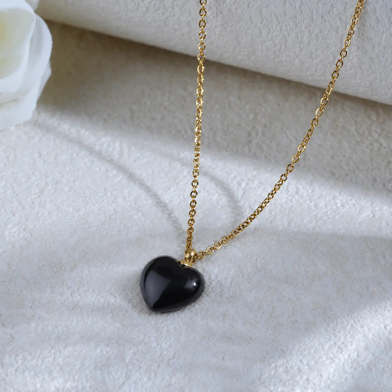 Flat lay aesthetic view of the MEHRZEB black heart gold necklace with a white rose prop and soft shadows.