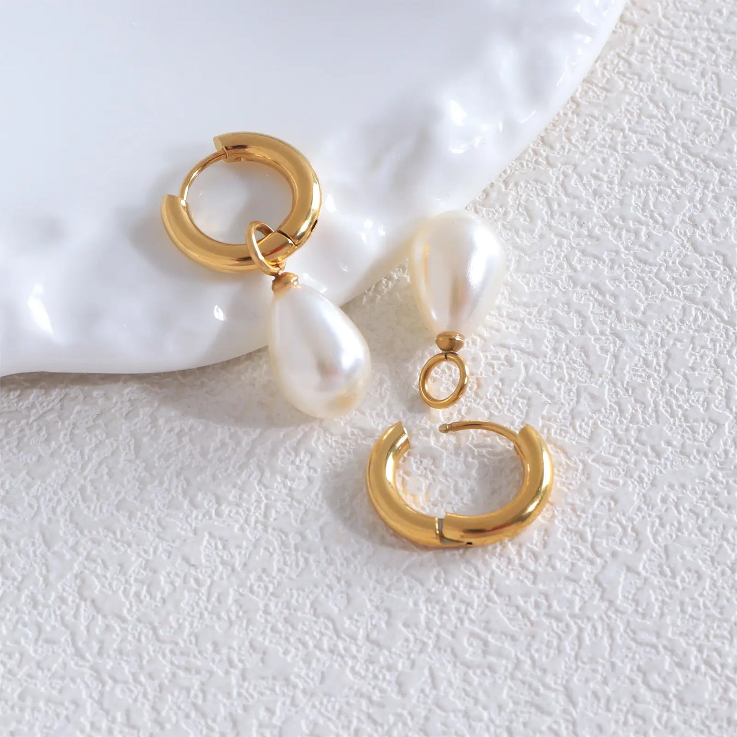 Flat lay aesthetic view of the MEHRZEB 18k gold pearl drop earrings on a textured white prop.
