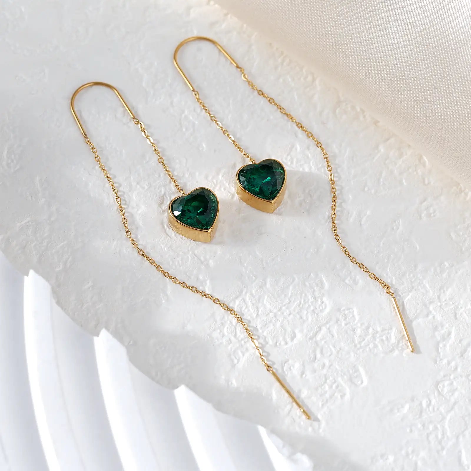 Flat lay aesthetic view of the MEHRZEB 18k gold threader earrings with green heart stones on a textured white prop.