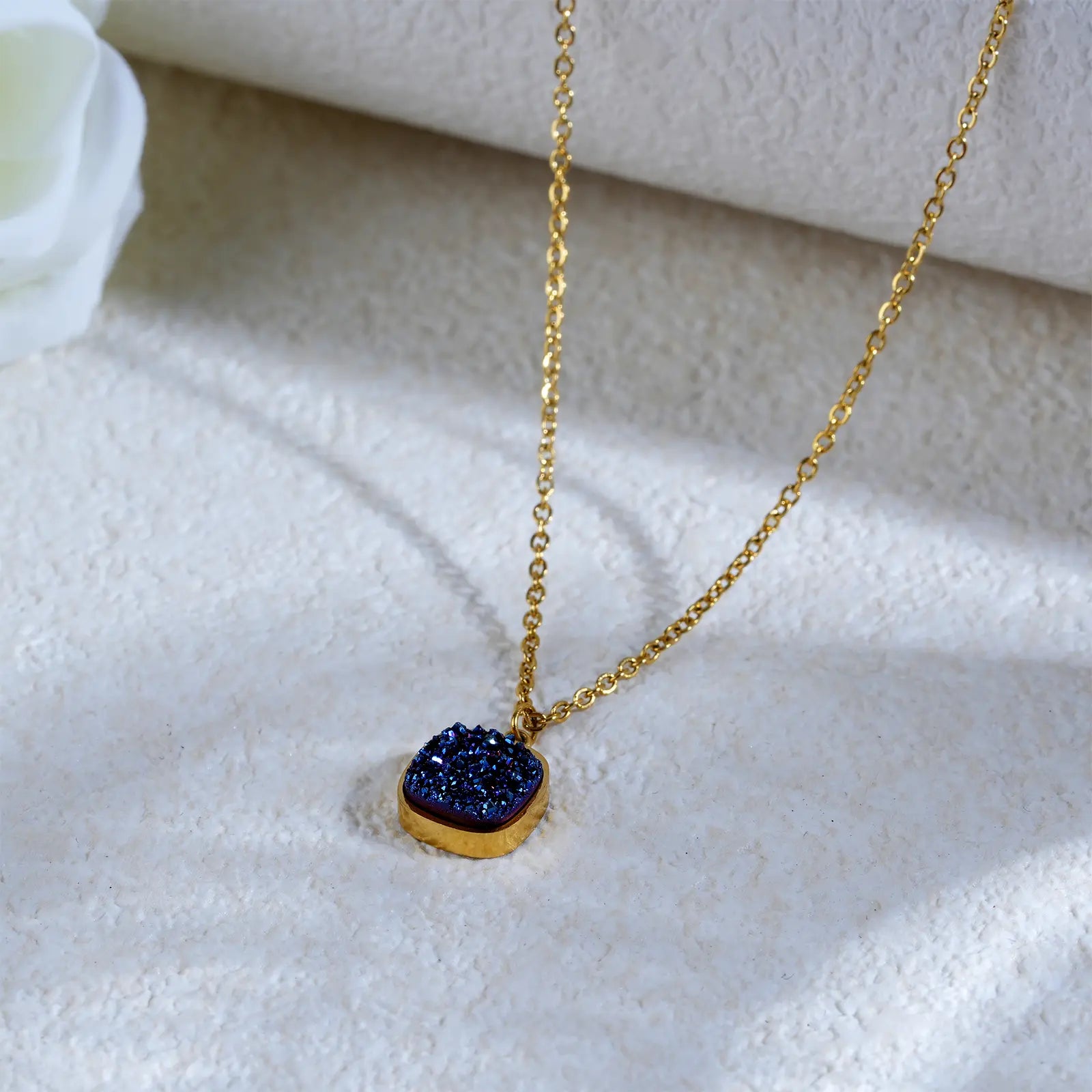 Elegant MEHRZEB gold plated blue pendant necklace displayed next to a white rose on a textured luxury background.