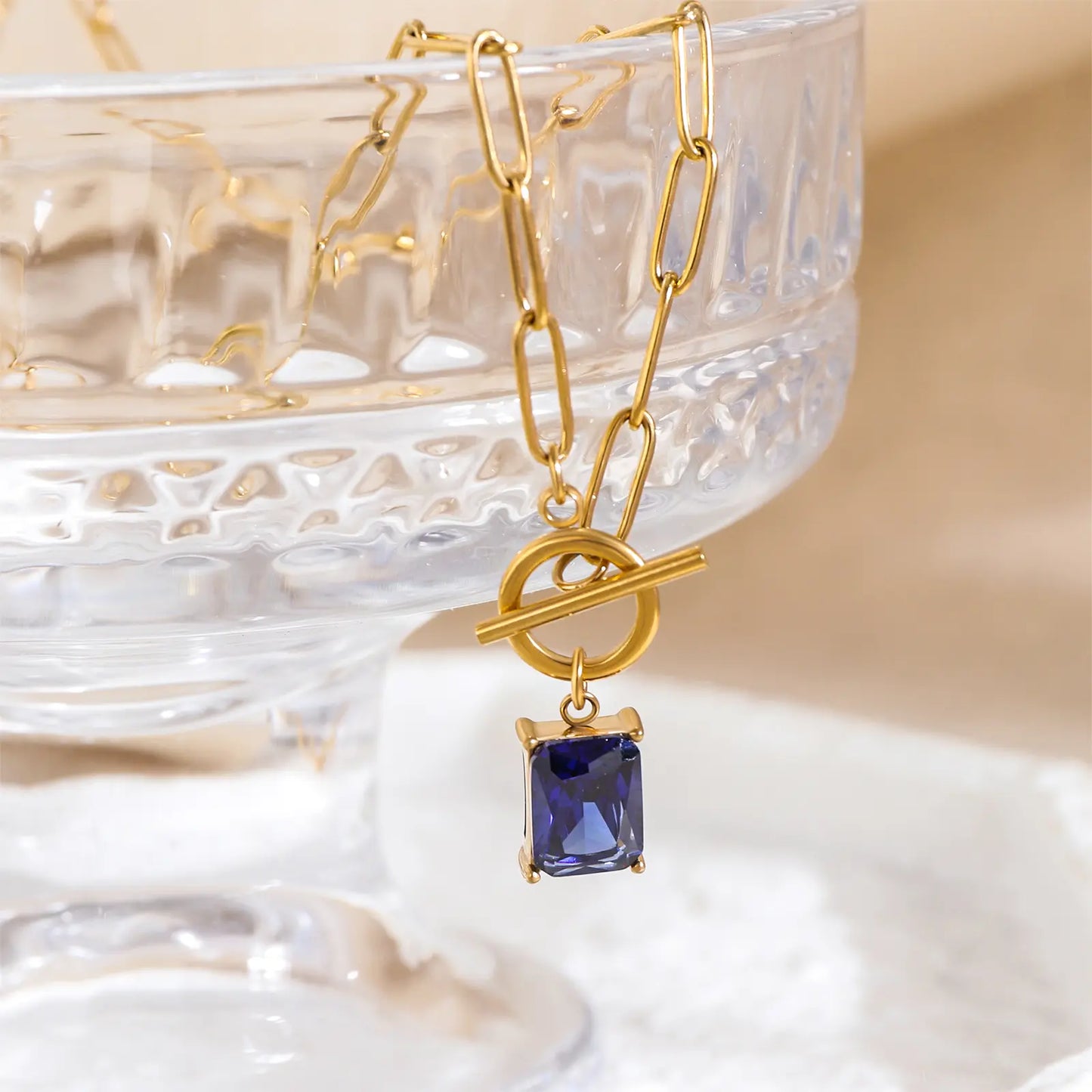 Close up of MEHRZEB blue zircon pendant and toggle clasp resting on a crystal glass prop.