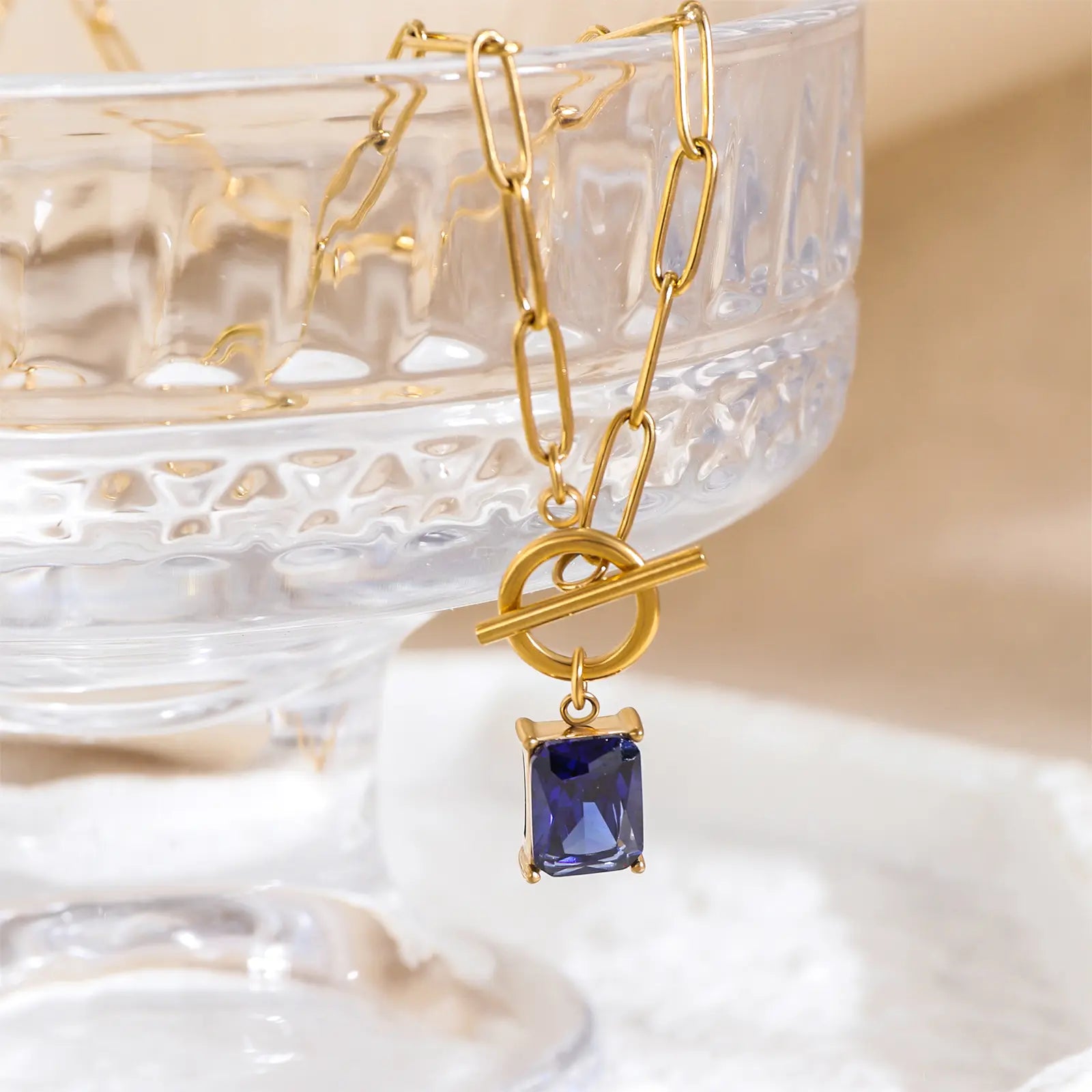 Close up of MEHRZEB blue zircon pendant and toggle clasp resting on a crystal glass prop.